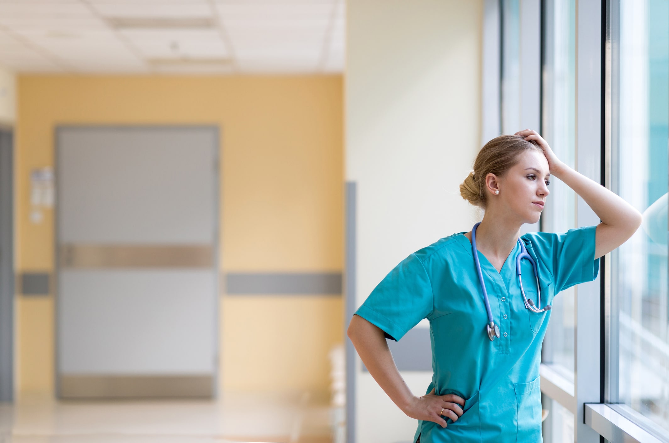 Tired female nurse standing in hospital corridor looking out of the window.